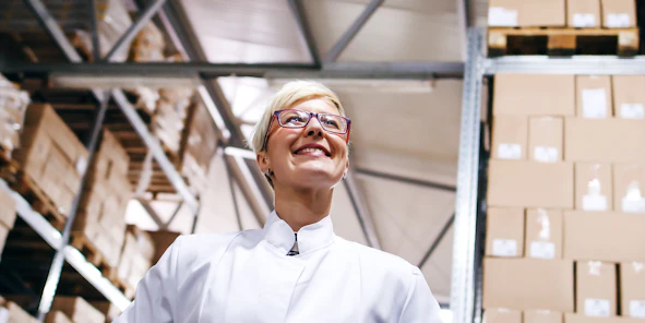 Eine Frau mit Brille steht lächelnd in einer Lagerhalle